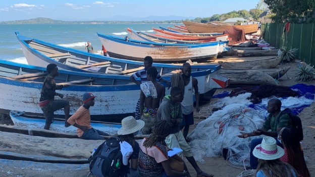 Small fish laid out to dry on raised racks along a sandy lakeshore, with a child standing nearby and water in the background.